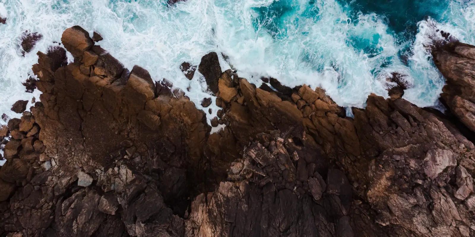 Aerial view of a rugged coastline with waves crashing against jagged rocks. The ocean is deep blue with white foam from the surf.